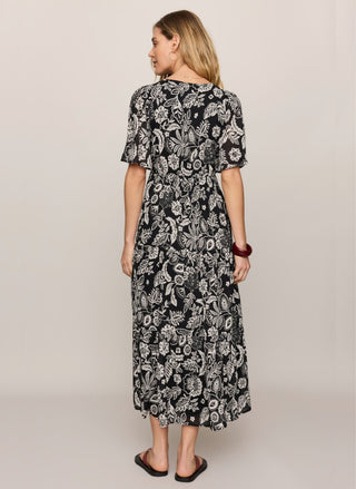 A woman shown from the back wearing a short-sleeve midi dress with a black and white floral paisley pattern, standing against a plain light beige studio background.