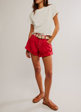 Front view of bright red high-rise denim shorts with raw hem, five-pocket styling, zip fly, and snug pull-on fit on a model’s hips.