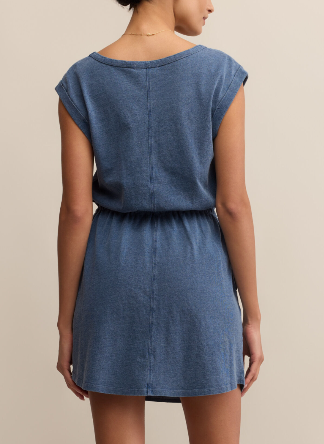 Woman wearing Z Supply Dayshift Denim Mini Dress in soft washed blue, featuring drawstring waist and cap sleeves, styled simply with gold jewelry.