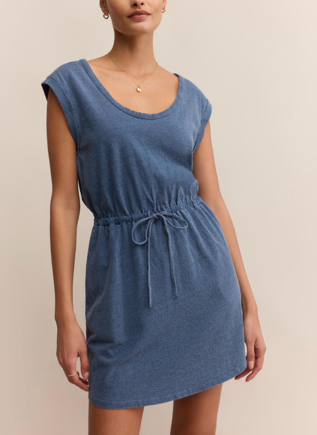 Woman wearing Z Supply Dayshift Denim Mini Dress in soft washed blue, featuring drawstring waist and cap sleeves, styled simply with gold jewelry.
