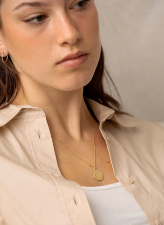 Woman wearing a gold necklace with a beige shirt and white top against a neutral background