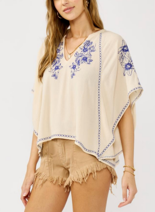 Woman wearing a cream top with blue floral embroidery on a plain background. 