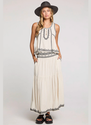 Woman wearing a long white dress with black patterns and a hat on a plain background