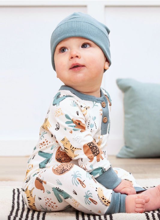 Baby wearing a patterned outfit and blue hat sitting on a couch.