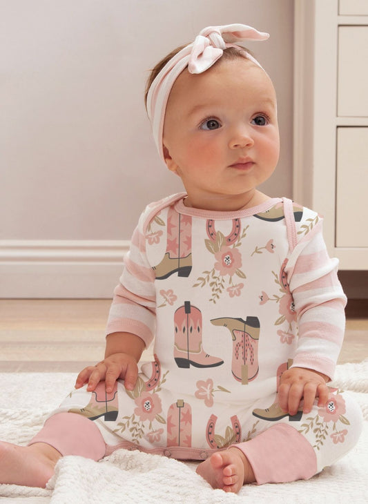 Baby wearing a onesie with cowboy boot and flower pattern, sitting on a light-colored floor.