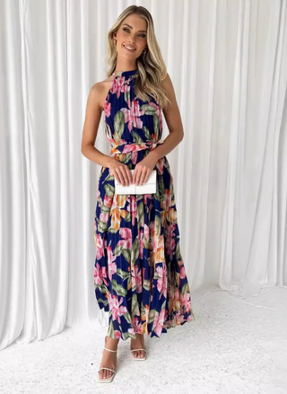 Model wearing the Midnight Blooms Pleated Halter Maxi Dress: a navy chiffon maxi with pintucked halter neckline, self-tie waist belt, and flowing pleated skirt featuring oversized fuchsia, blush, and olive floral print; styled with white mini clutch and white strappy sandals.