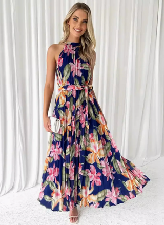 Model wearing the Midnight Blooms Pleated Halter Maxi Dress: a navy chiffon maxi with pintucked halter neckline, self-tie waist belt, and flowing pleated skirt featuring oversized fuchsia, blush, and olive floral print; styled with white mini clutch and white strappy sandals.
