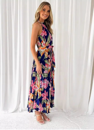Model wearing the Midnight Blooms Pleated Halter Maxi Dress: a navy chiffon maxi with pintucked halter neckline, self-tie waist belt, and flowing pleated skirt featuring oversized fuchsia, blush, and olive floral print; styled with white mini clutch and white strappy sandals.