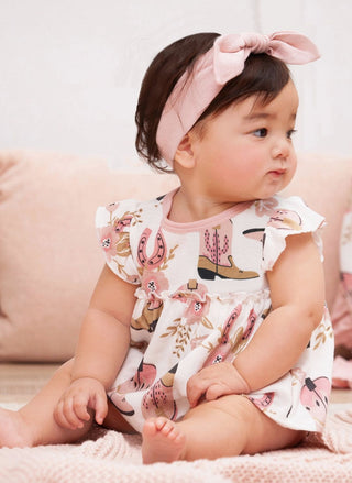 Baby wearing a floral romper with cowboy boots and pink headband on a soft surface