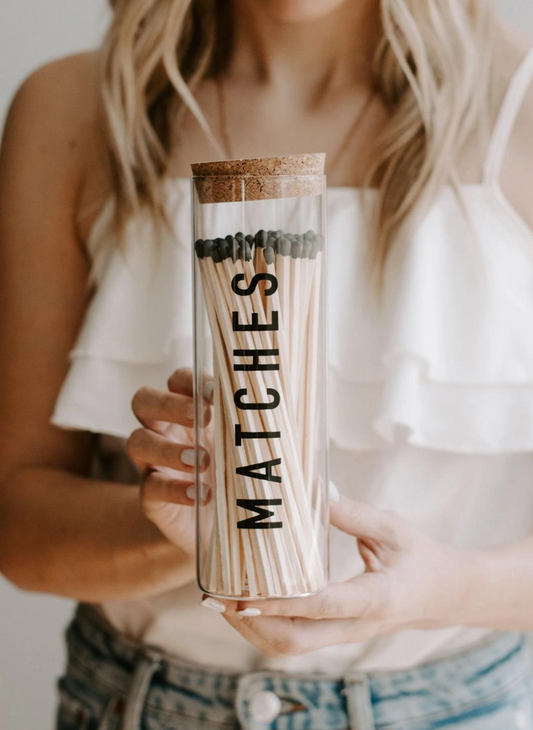 Model holding product.  Tall glass round canister for 7" matches.  Front of glass jar says "MATCHES" in black lettering.  Cork top.