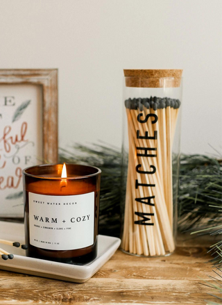 Image of matches sitting on a wood shelf with a burning candle and artwork behind it.