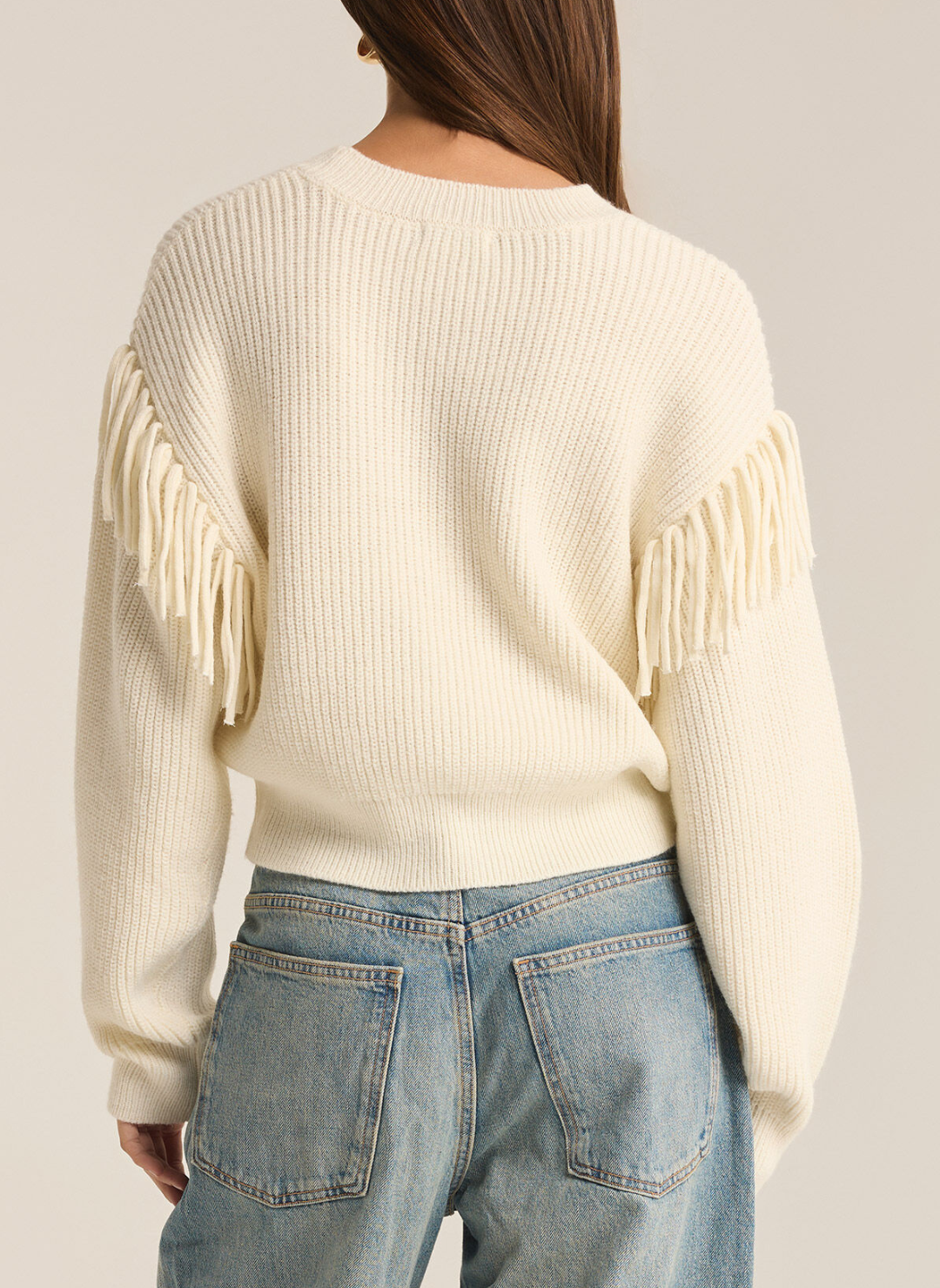 Model is wearing On the Fringe sweater paired with denim jeans displaying the detailed fringe on sleeves. Back view with white background.