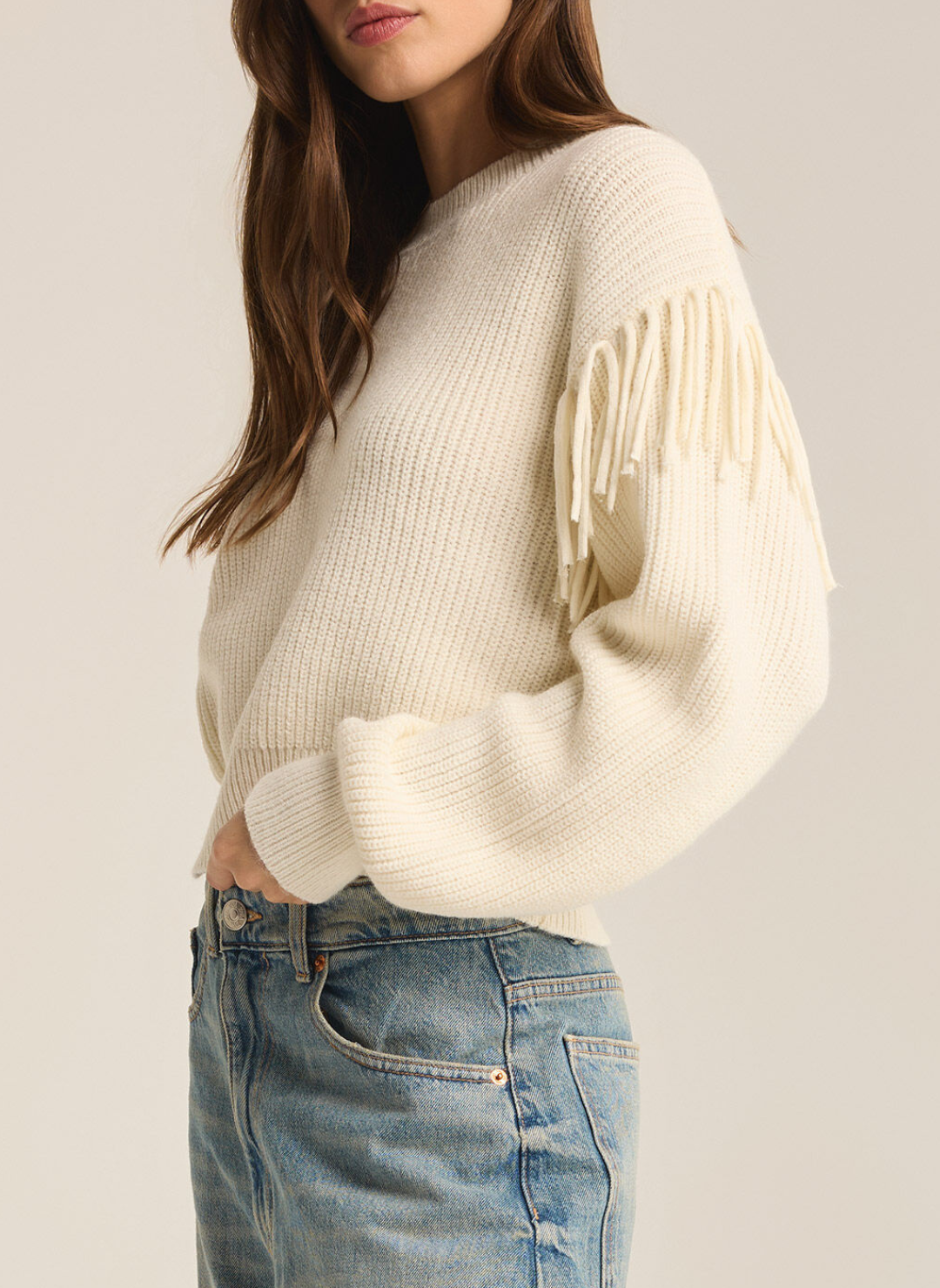 Model is wearing On the Fringe sweater paired with denim jeans displaying the detailed fringe on sleeves. Side view with white background.