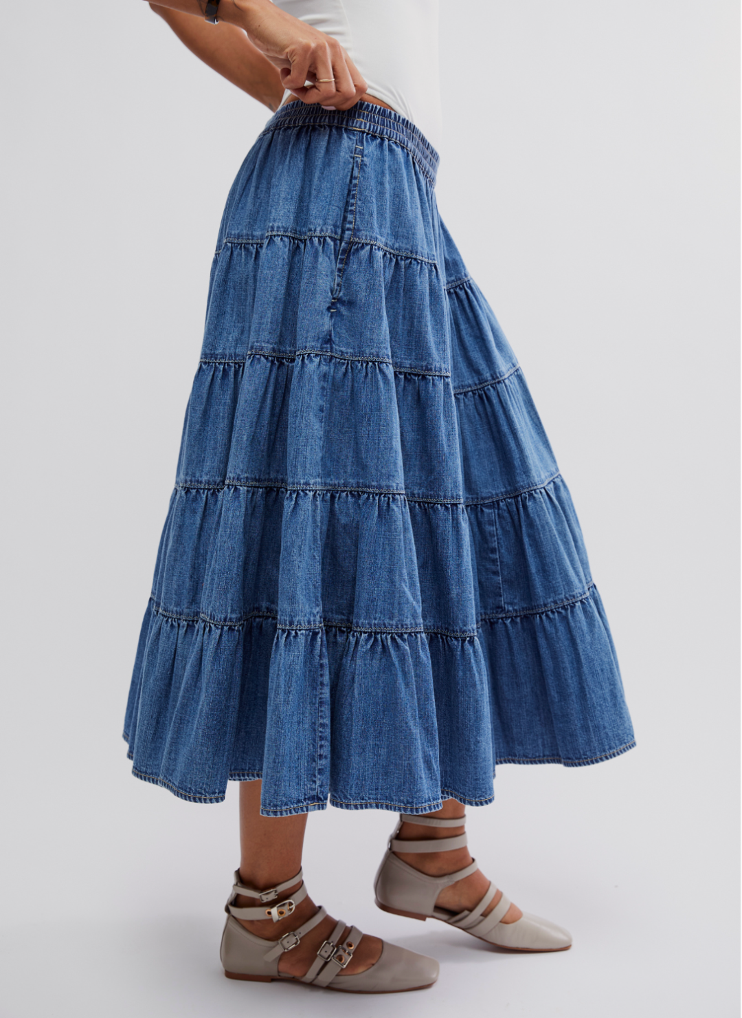 Side view of the Full Swing Denim Skirt. Model is standing in front of a white background and is wearing a white body suit with gray shoes.