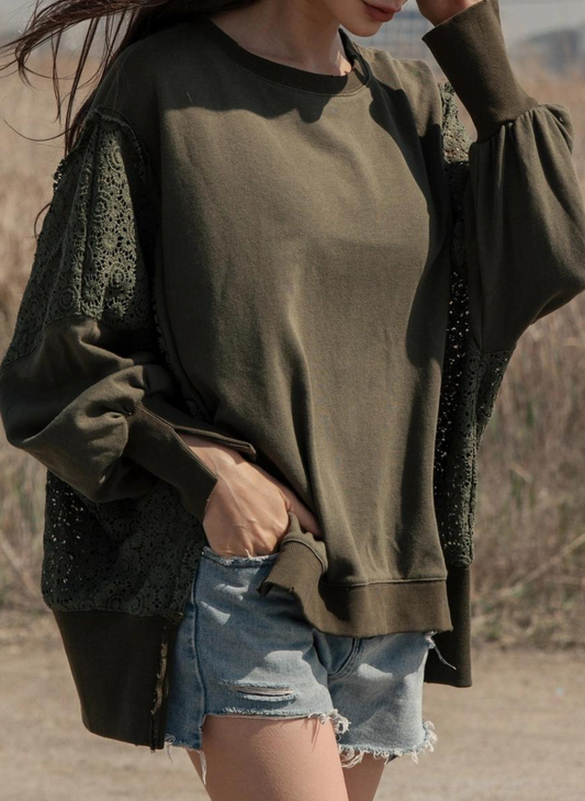 Image of model with left hand to her face, an oversized sweatshirt in an olive green color.  Sweatshirt has a crew neck, lace insets and side slits at the side seam and at the cuff.