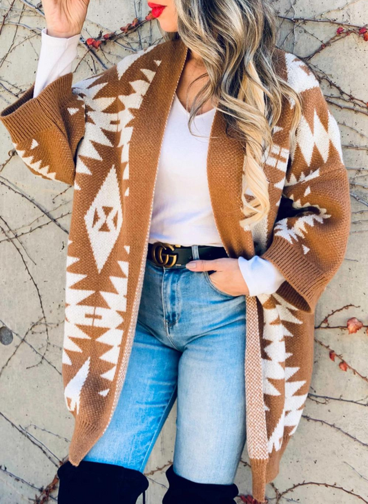 Model is wearing a caramel open front Aztec printed knitted cardigan with a white shirt underneath, black belt, and blue jeans with models hand in her pocket.