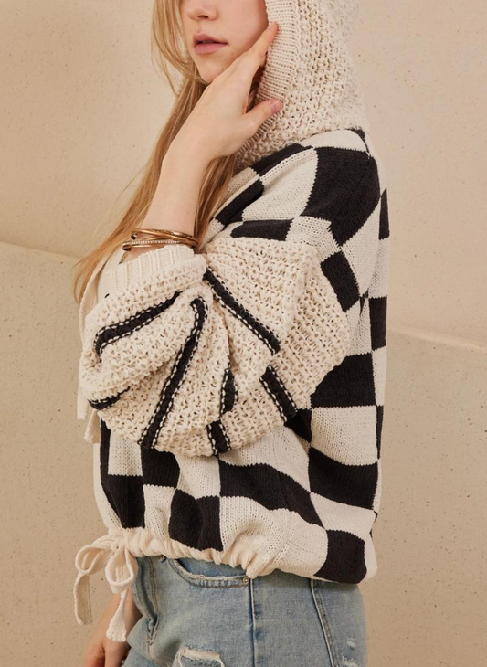 The model is posing with her left arm on her face, wearing a black and off white sweater. The sweater is checkered print, with a fun stripe design in the sleeves, and a cute drawstring bow tie.