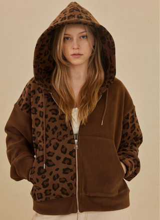 Model wearing Lioness Leopard Zip Up Hooded Sweatshirt in brown with black leopard print with the hood over her head and hands in the pockets.