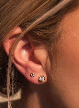 Close-up of a model's ear wearing petite gold FYB Marisol Bezel Studs 3mm with sparkling round clear stones, styled alongside additional delicate earrings.
