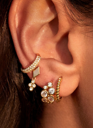 Close-up of a model's ear wearing the gold FYB Tory Ear Cuff with sparkling clear stones, styled alongside gold hoops and dangling earrings.